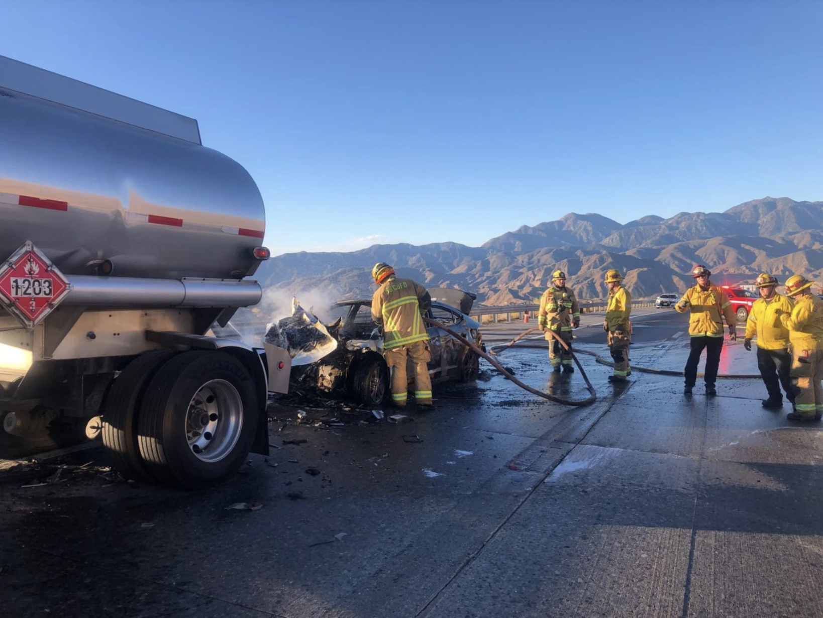 An image showing a roadside vehicle accident where a small passenger car has driven into the rear end of a heavy freight tanker truck. Around the passenger car are firefighters working to extinguish the flames from the front of the vehicle with a fire hose.