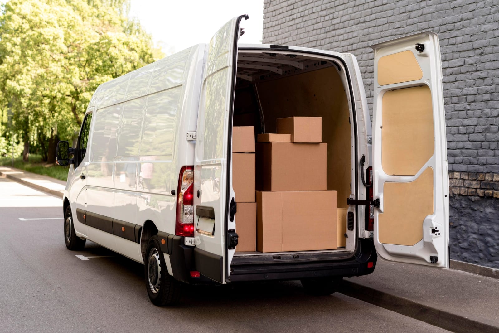 An image of a white panel van parked alongside a roadway. The rear doors of the van are open with multiple cardboard boxes stacked inside indicating that it is a delivery van.